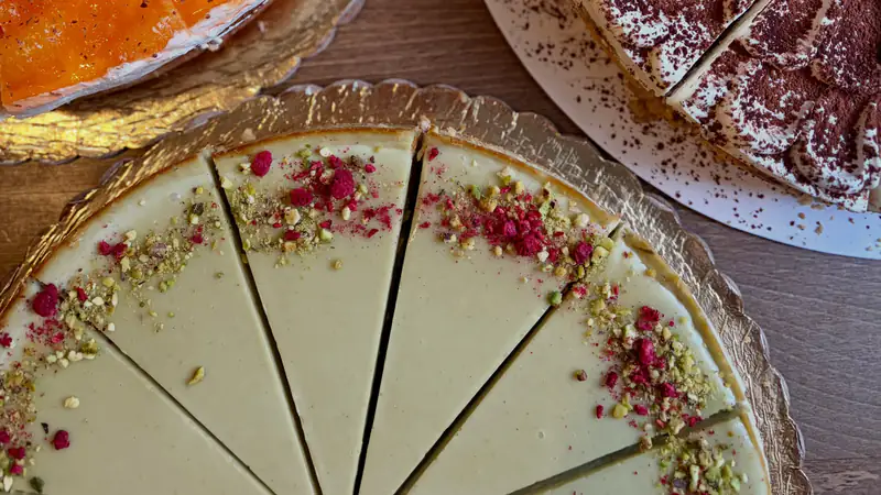 Nakrájaný pistáciový cheesecake s drvenými pistáciami a ružovou posýpkou, vedľa neho mandarínková torta a tiramisu cheesecake, pohľad zhora